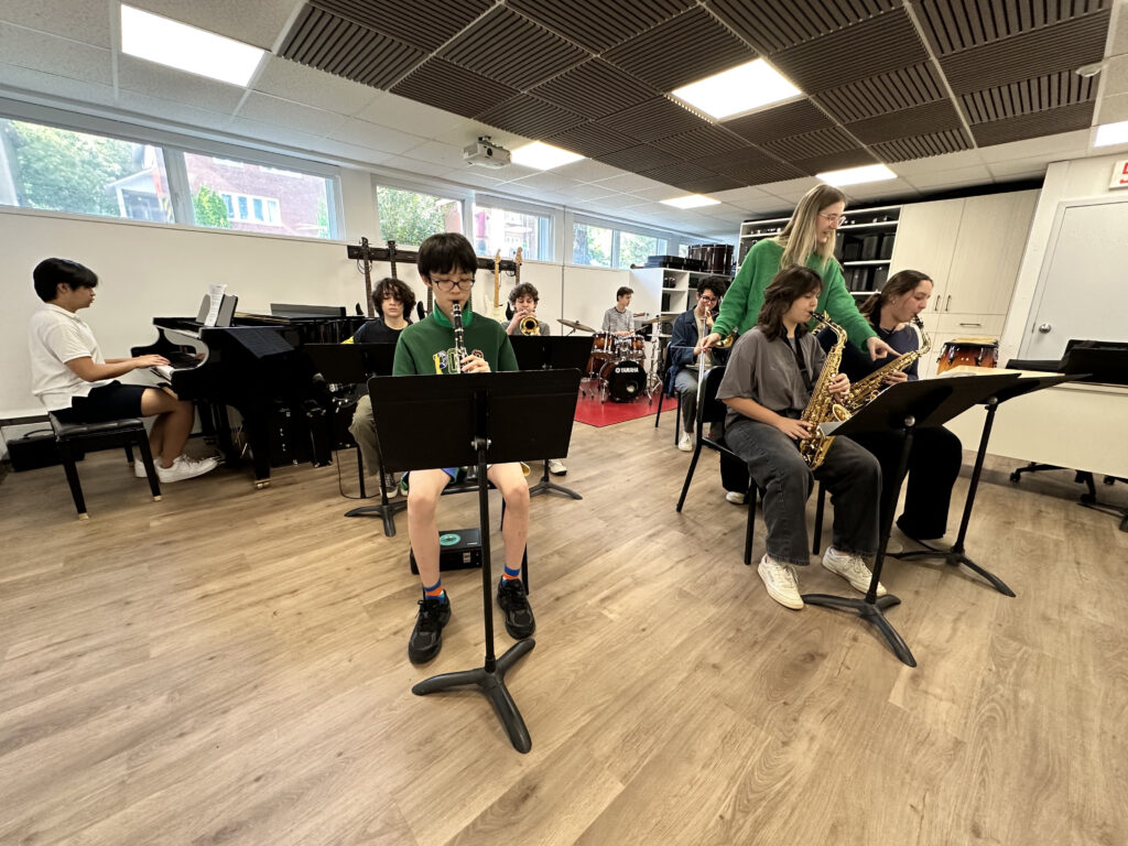 A group of high school students play instruments together in a bright music studio. A teacher stands beside two saxophone players, offering guidance. Other students play piano, trumpet, drums, and clarinet. The room has wood floors, acoustic ceiling panels, and large windows that let in natural light.