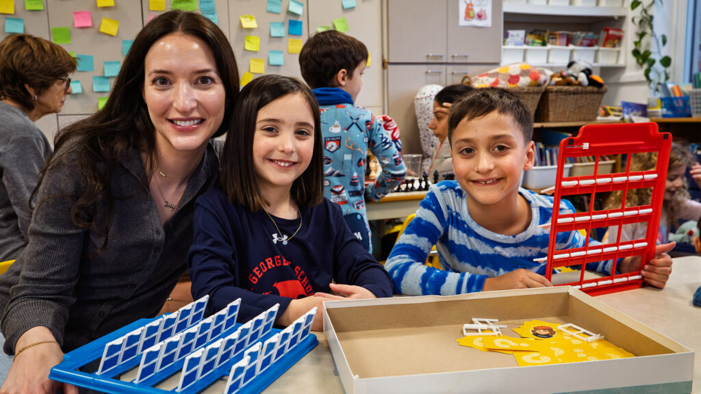 A boy and girls sit at a table in school. Beside them is a smiling woman with brown hair. There are flash card games on the table.