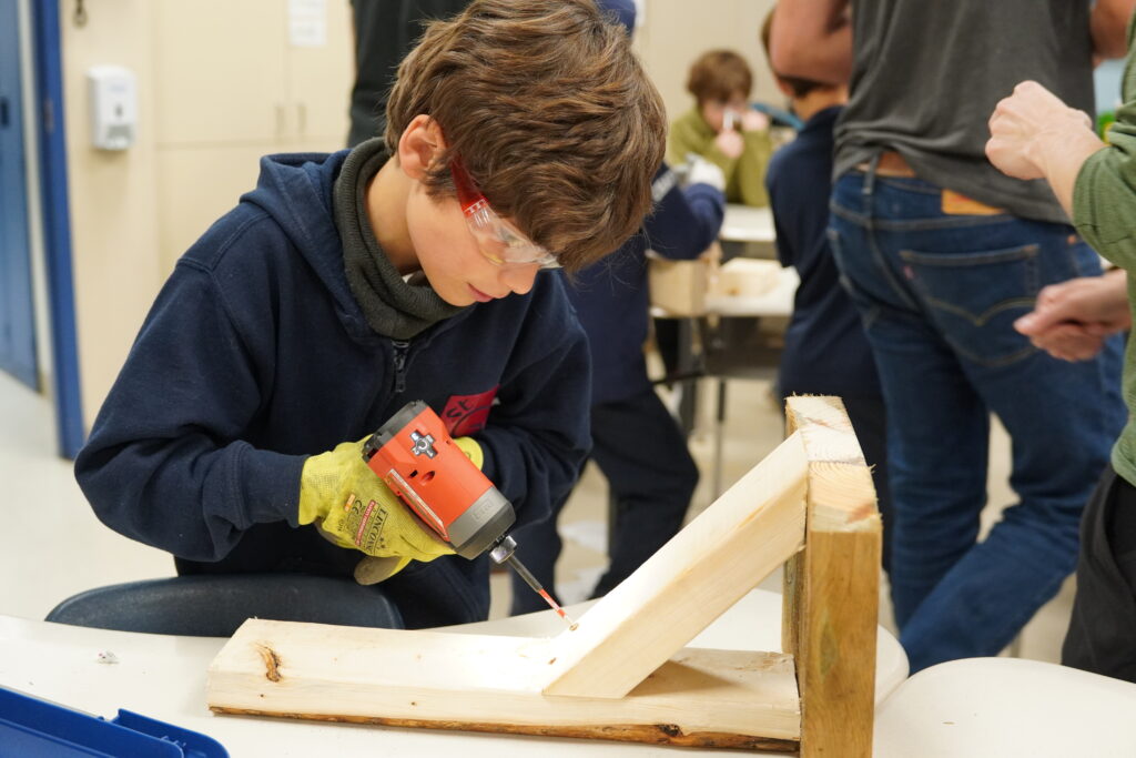 Elementary Student drills a piece of wood. He wears protective glasses.