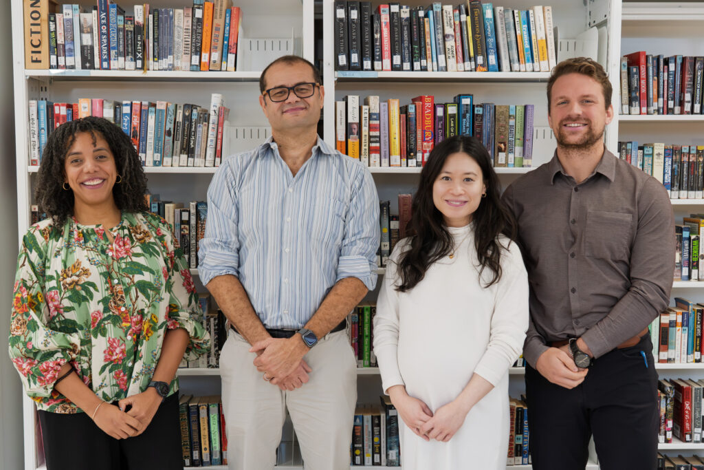 Four adults stand in front of a bookshelf. They are all of different nationalitiers. Two women and two men.
