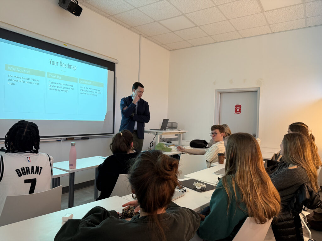 A man stands at the front of a class with a smart board behind him.