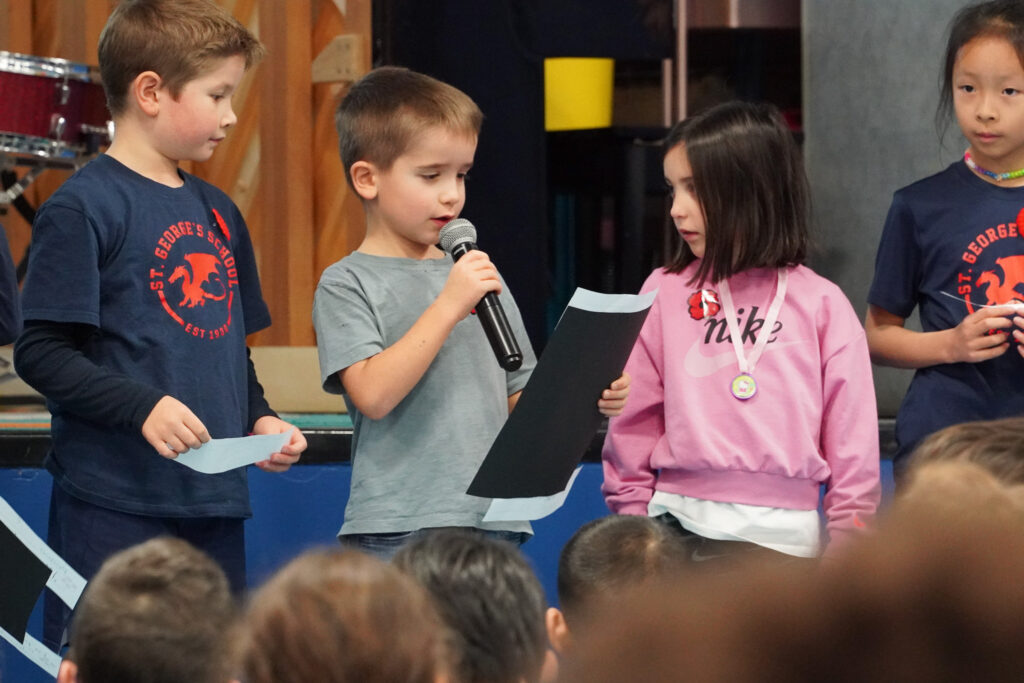 A group of young students stand in front of a stage. The boy holds a microphone and speaks into it.