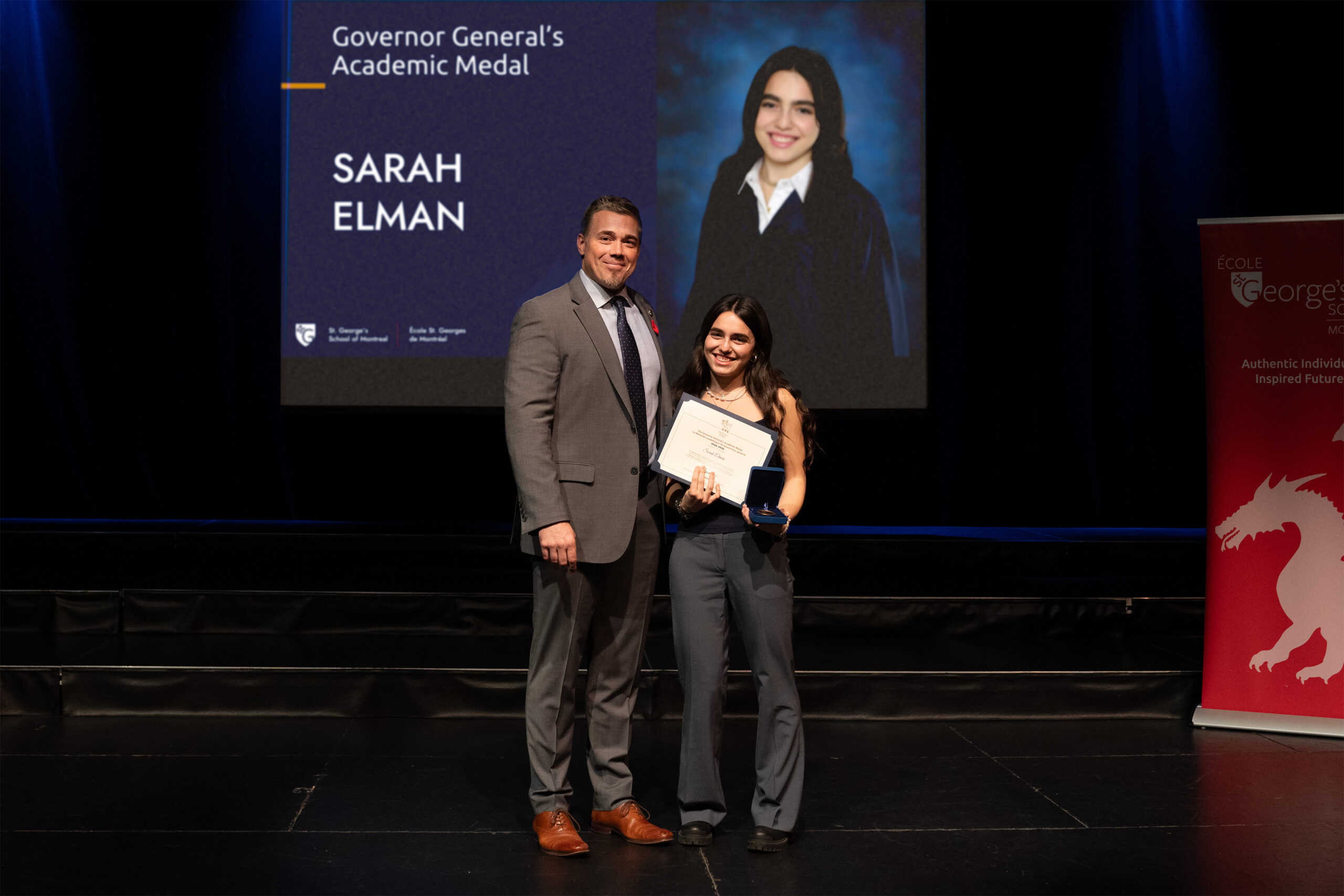A man and a high school female student stand on stage. The student is holding a certificate. Behind them is a screen projecting the student's photo.
