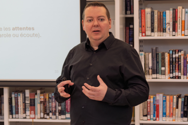 A man gestures with his hands in front of shelves of books.