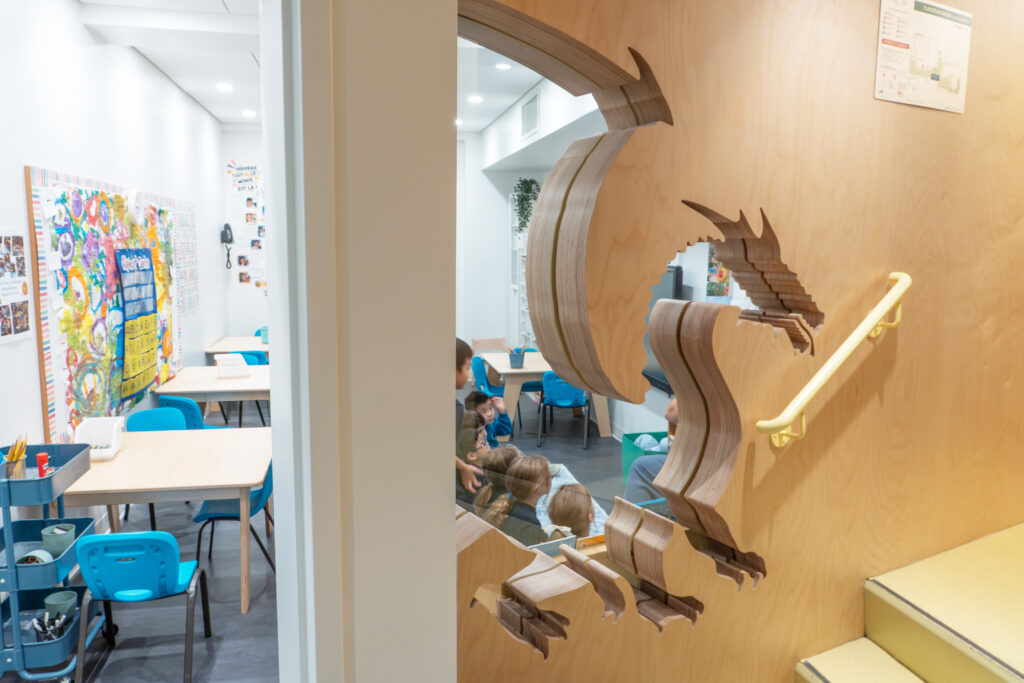 A cut out of a dragon silhouette window with wood frame. Looking through kindergarten children learn in class seated on the floor.