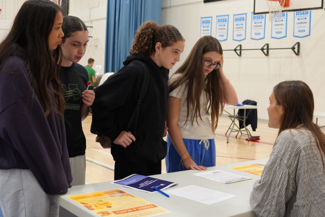 Female students stand at a table and speak to a woman seated behind it. They are in a gymnasium.