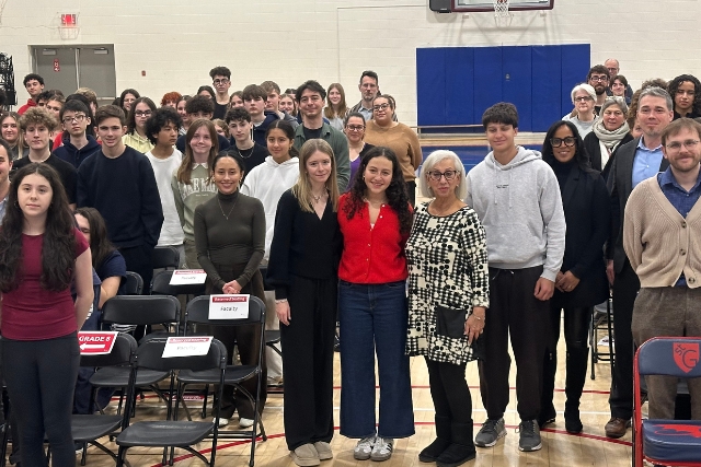 A crowd of high school students stand together with an older woman in a high school gym.