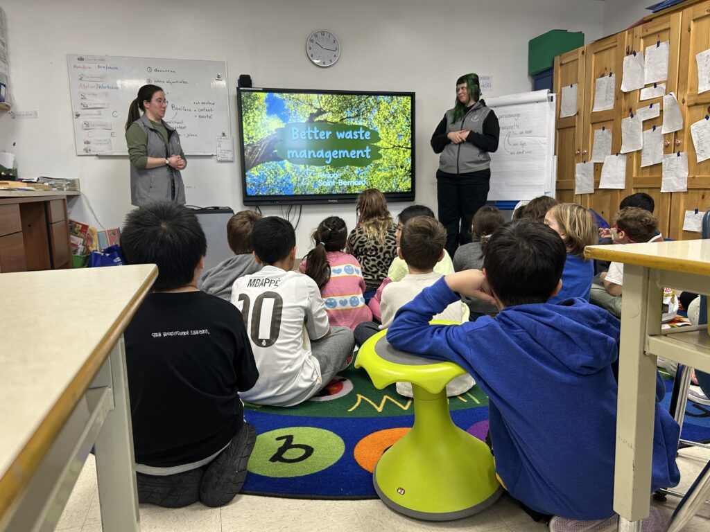 Students sit on the floor of a classroom and listen to a lecture by a woman in front of a screen.