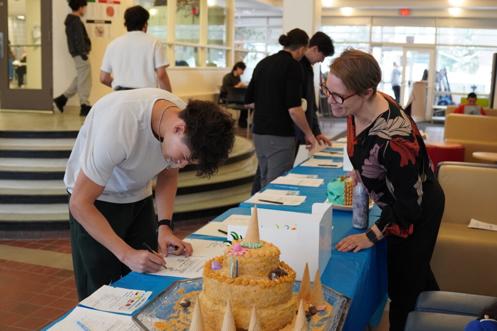 A student signs a sheet of paper. A woman is in front of him and a cake is on the table.