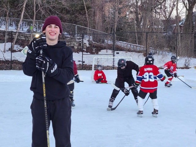 A teenage boy stands in the ice with a hockey sticks. More players are in the background.