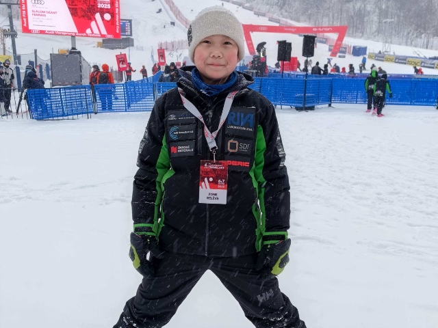 An asian boy skier stands at the bottom of a hill.