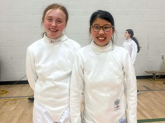 Two female fencers stand shoulder to shoulder.