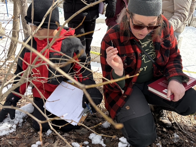 An elementary student and instructor kneeling in the snow to study winter animal tracks and buds as part of the Coyote Nature Program.