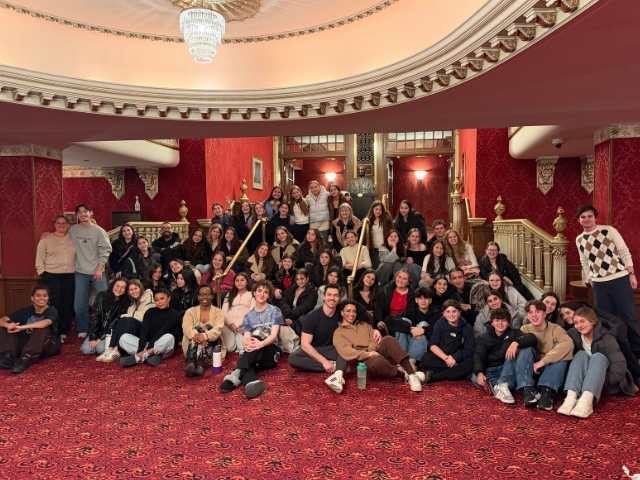 A group of G Major show choir students and staff posing for a large group photo on a red carpeted staircase in a theater lobby with alum Patrick Park ’12.