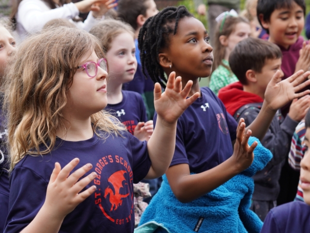 A group of young Elementary School students in St. George's t-shirts dance outdoors, arms raised and faces lifted, surrounded by classmates