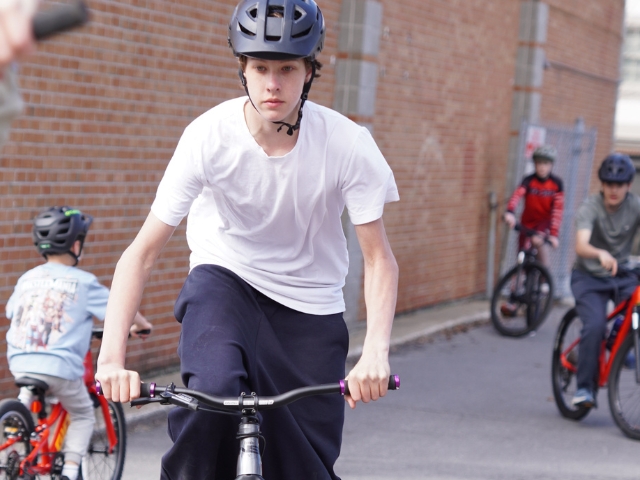 A teenage student in a white t-shirt and helmet rides a bike toward the camera in an outdoor school setting, with younger students on bikes visible in the background.