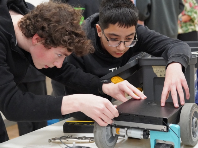 Two male students lean over a workbench, focused intently on assembling the mechanical components of a small robot.