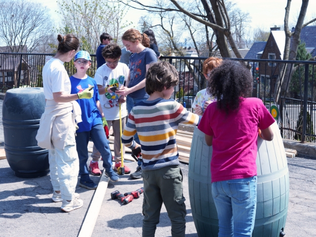 A group of Elementary School students gather outdoors on a rooftop garden space, building rain barrels together with lumber and tools nearby, guided by a teacher on a sunny day.