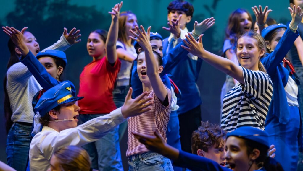 A group of teenagers sing and raise their hands. Some of them wear what looks like pilot and stweardess outfits.