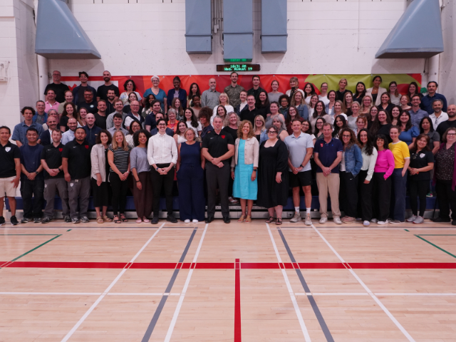 A large group of school staff members stand together for a group photo in a gymnasium with red and green wall padding and a scoreboard in the background.