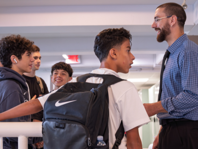 A teacher in a blue checked shirt and tie smiles while talking with a group of teenage boys in a school hallway; one boy in the foreground wears a black Nike backpack.