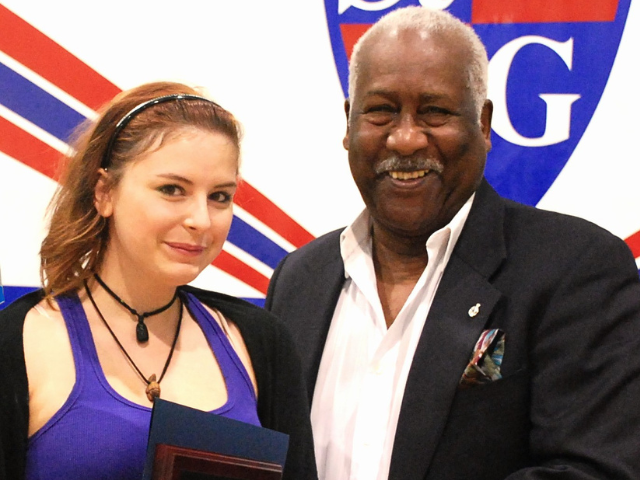 A young white woman stands with an older black gentleman in front of a logo with a G in it. They are both smiling.