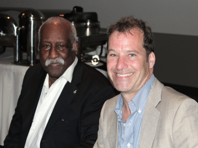 A white man in a beige suit jacket sits with an older black man in a black suit jacket. They are both smiling.