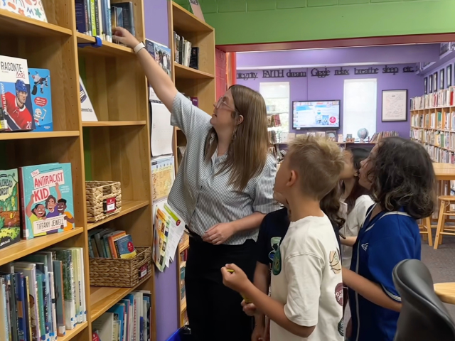 A woman reaches up to get a book from the shelf for students.
