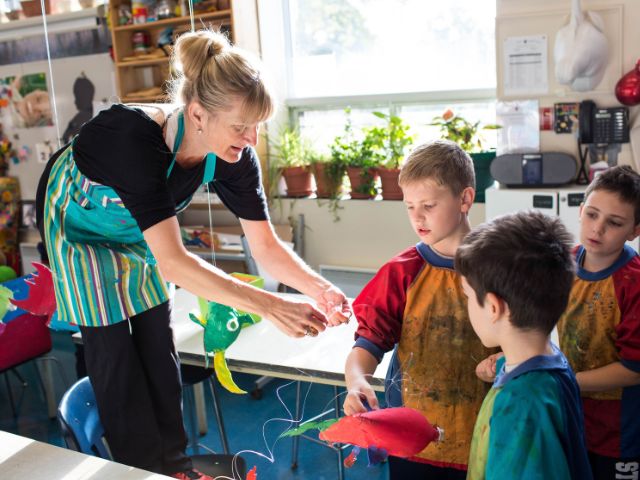 A teacher in a striped apron holds up a hanging sculpture of colourful fish as three young students in paint-splattered smocks watch closely in a sunny art room.