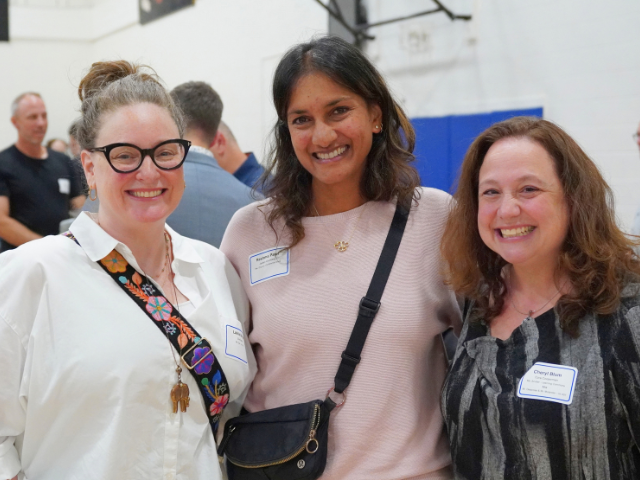 Three smiling women wearing name tags stand together in a busy school gymnasium.