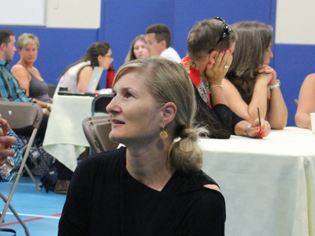 A woman with her hair tied back looks toward the front of a gymnasium where adults sit at tables covered in white tablecloths.