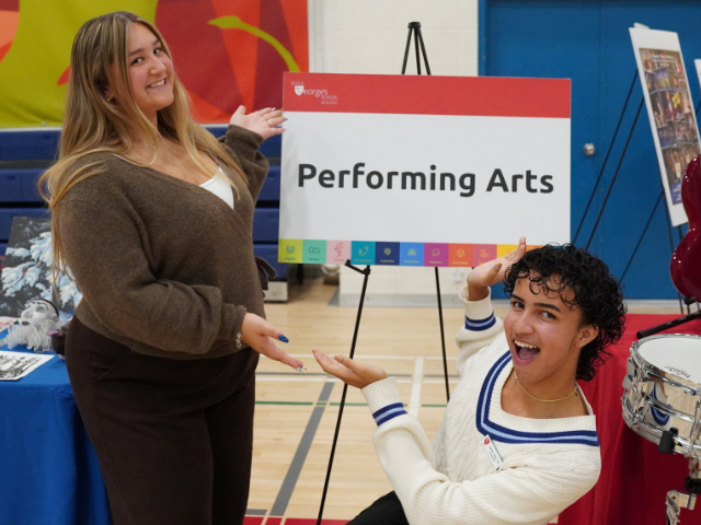 Two smiling students gesture toward a Performing Arts sign at a display table.