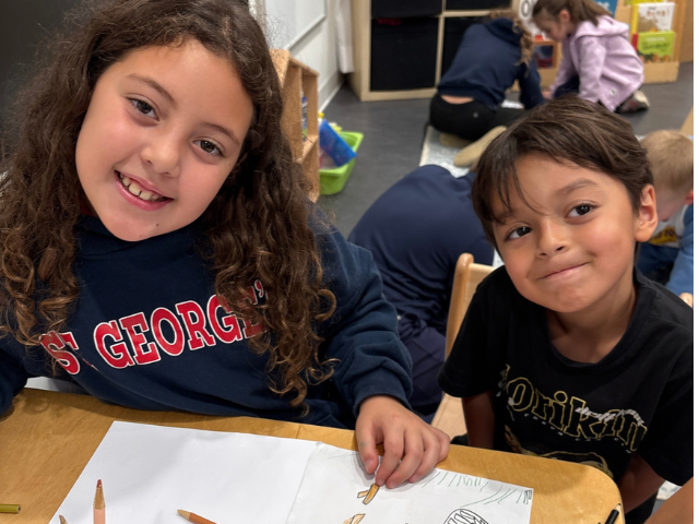 An older student in a St. George's sweatshirt and a younger student smile at the camera while working on a sheet of paper at a classroom table.