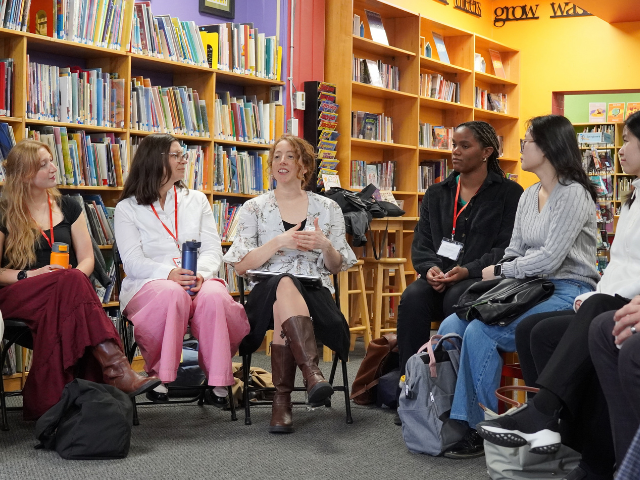 group of adults sits in a circle of chairs in a colourful school library lined with bookshelves, listening as one person speaks.