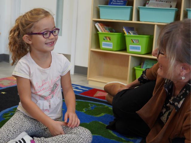 A young student sitting on a classroom rug smiles at a teacher seated beside her