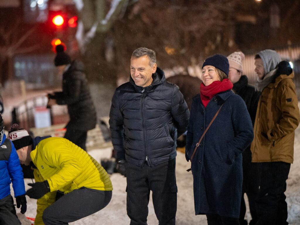 Parents in winter coats and scarves smile and chat together outdoors on a snowy evening, with other families gathered around them.