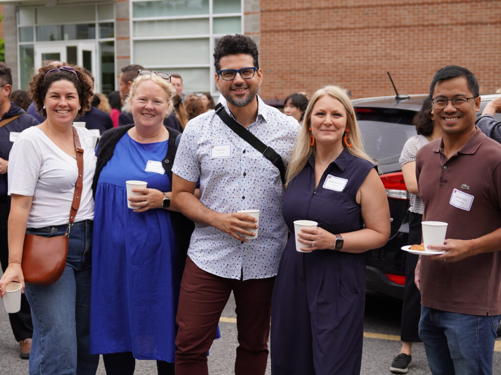 Four adults smile together outside a brick school building, holding coffee cups, with other parents gathered behind them.