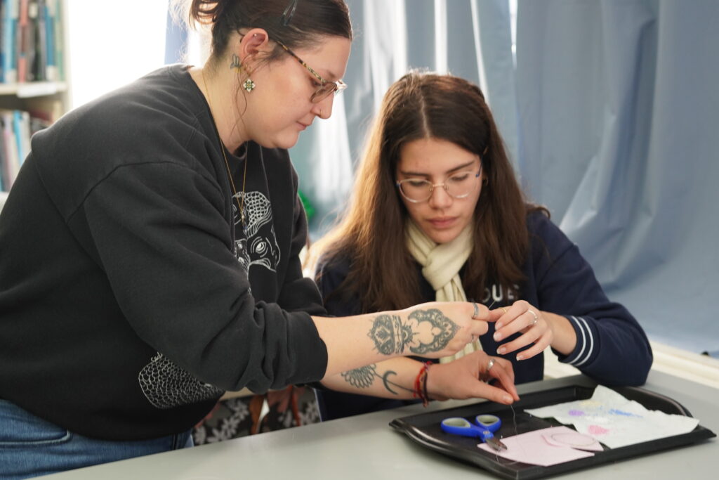 An artist leans over a table to help a student thread a needle. A tray in front of them holds scissors, fabric and a small beading project.