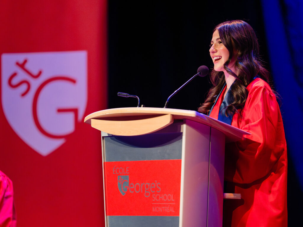 A smiling graduate in a red gown and navy stole speaks into a microphone at a podium bearing the St. George’s School of Montreal logo, with a large red banner of the school crest behind her.