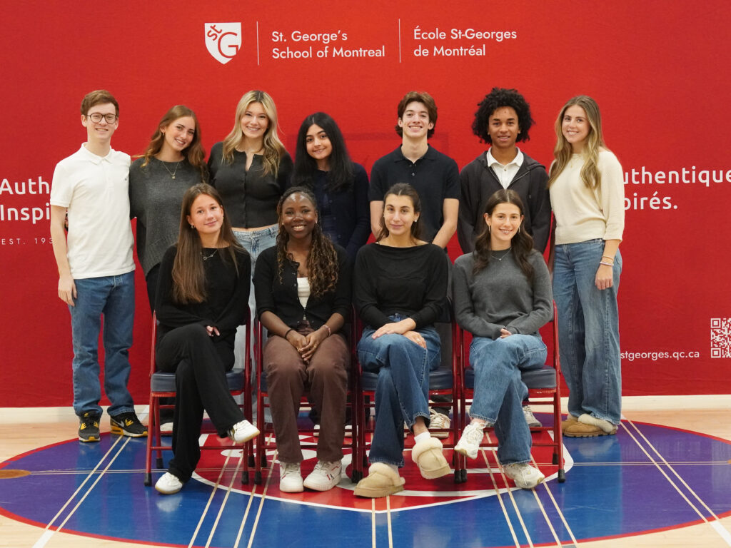 Grade 12 students smile in two rows, four seated on red chairs and seven standing, in front of a red St. George’s School of Montreal banner on the school gym floor.