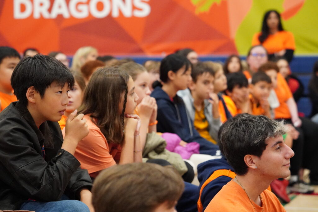 Students of different ages, many wearing orange shirts, sit on the gym floor and listen attentively beneath a large Dragons banner.