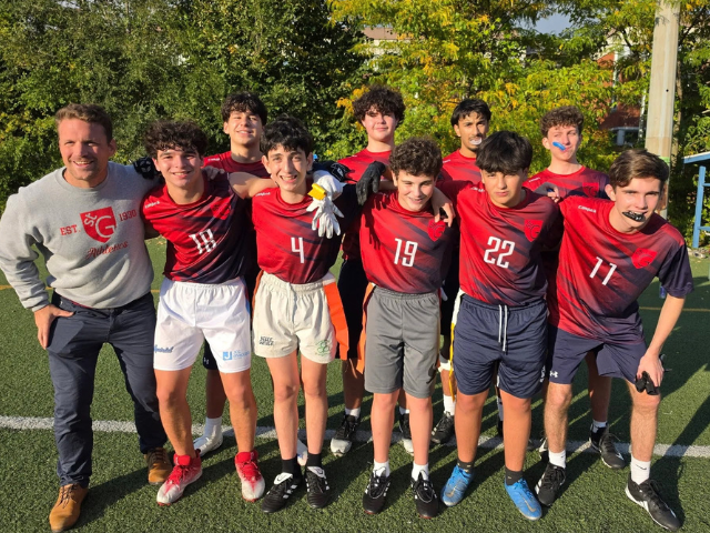 A group of smiling student athletes in red and navy jerseys stands with their arms around one another on a turf field, joined by an adult in a grey St. George's sweatshirt.