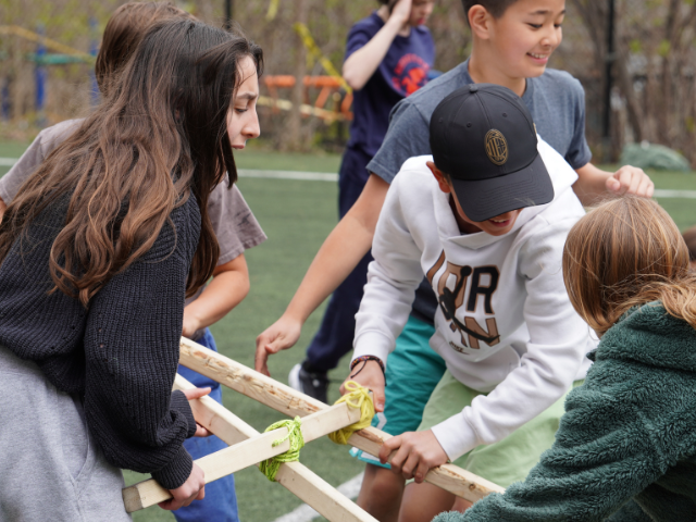 A group of students on a turf field grip a wooden frame lashed together with bright yellow rope, smiling and concentrating as they lift it.